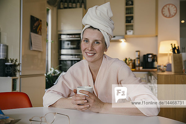 Porträt einer lächelnden Frau mit Handtuchturban  die mit einer Tasse Kaffee bei Tisch in der Küche sitzt