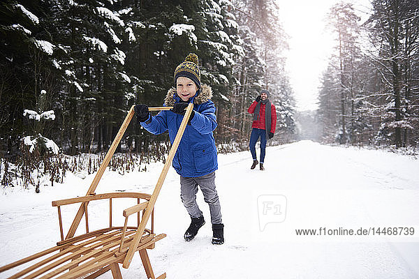Little boy pushing sledge in winter forest while his father watching him from background