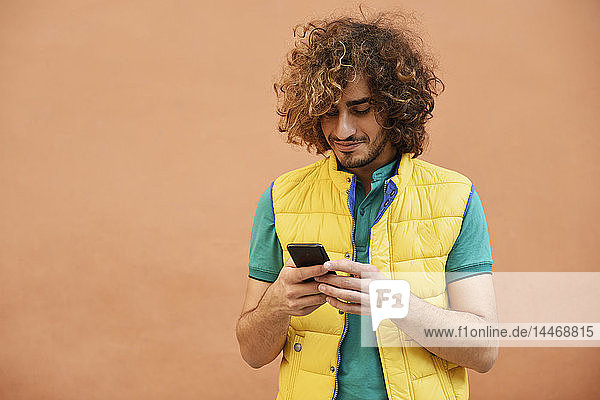 Smiling young man with curly hair wearing yellow waistcoat looking at cell phone