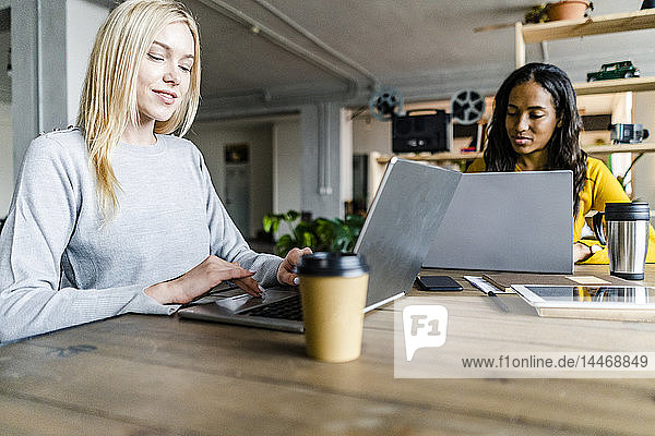 Two young businesswomen sitting at conference table in loft office using laptops