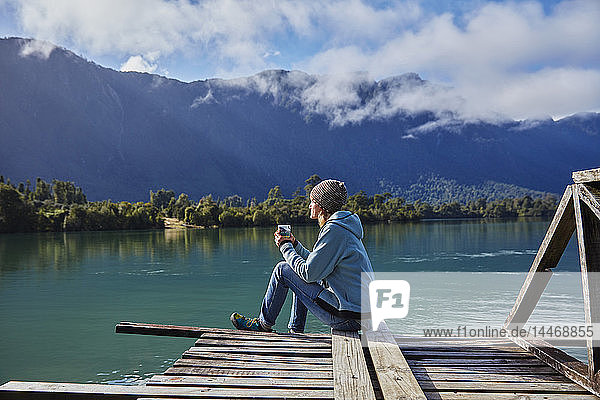 Chile  Chaiten  Lago Rosselot  Frau sitzt auf dem Steg und hält einen Becher