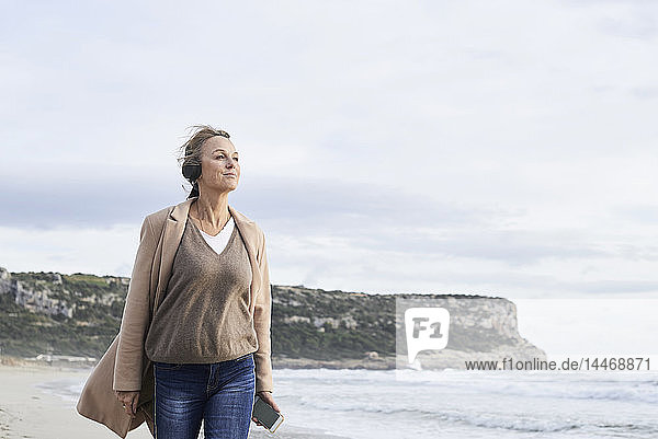 Spanien  Menorca  ältere Frau mit Smartphone und drahtlosen Kopfhörern am Strand im Winter