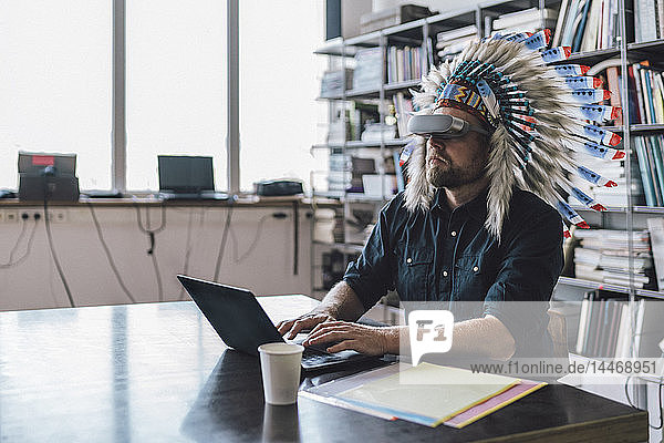 Man wearing Indian headdress and VR glasses in office  using laptop