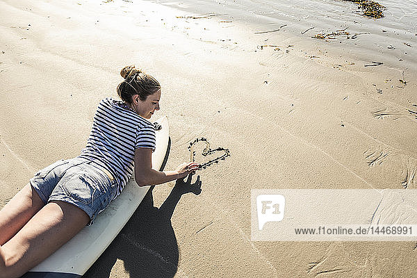 Junge Frau mit Surfbrett  die ein Herz in den Sand zeichnet