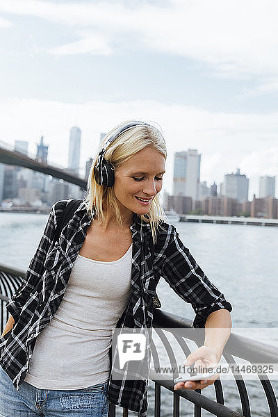 USA  New York City  Brooklyn  lächelnde junge Frau steht mit Kopfhörern und Handy am Wasser