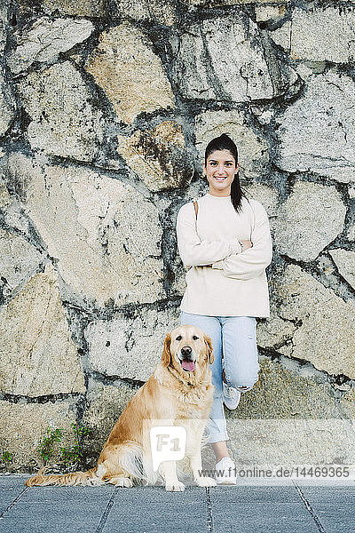 Porträt einer lächelnden jungen Frau mit ihrem Golden-Retriever-Hund an einer Steinmauer