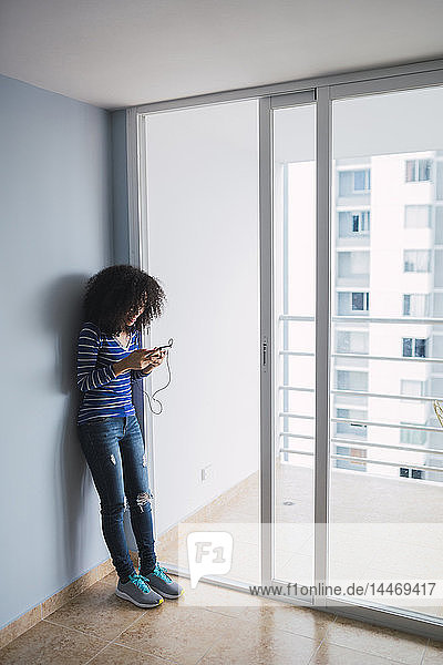 Lächelnde junge Frau steht an der geöffneten Balkontür ihrer neuen Wohnung und benutzt ein Smartphone