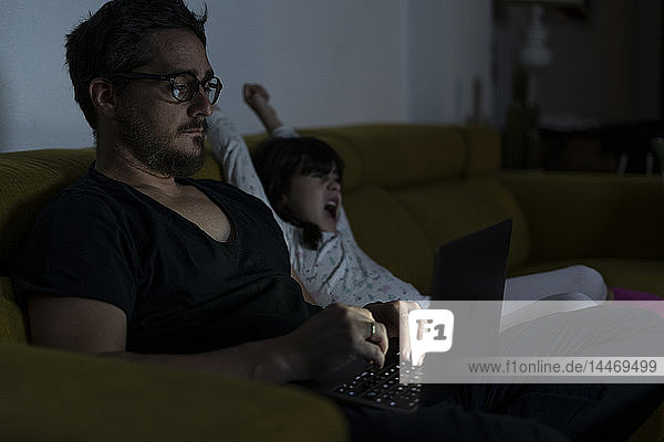 Father with daughter sitting on couch using laptop at night