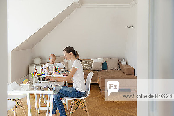 Mother using laptop and little daughter playing at table at home