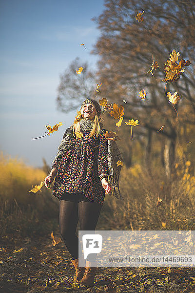 Glückliche schwangere Frau steht auf Waldweg und spielt mit Herbstlaub