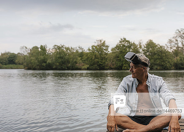 Älterer Mann  der mit einer VR-Brille auf einem Steg an einem See sitzt