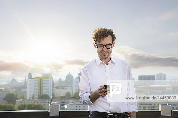 Deutschland  Berlin  lächelnder Geschäftsmann auf der Dachterrasse beim Blick auf das Smartphone am Abend