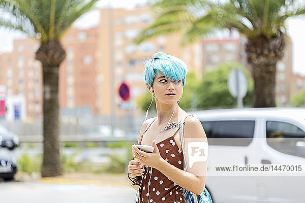Spanien  Porträt einer jungen Frau mit blau gefärbten Haaren  die mit Kopfhörern und Smartphone Musik hört
