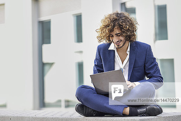 Smiling young businessman with curly hair wearing blue suit sitting on bench outdoors using laptop