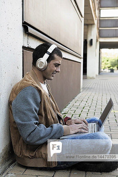Young man sitting outdoors wearing headphones and using laptop