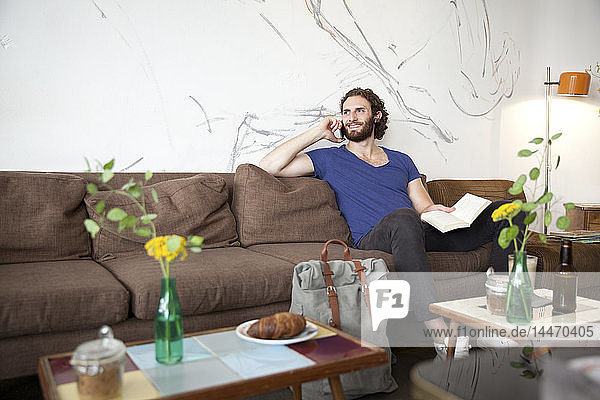 Porträt eines jungen Mannes am Telefon  der in einem Café mit Buch auf der Couch sitzt