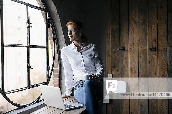 Smiling businesswoman with laptop and cup of coffee looking out of window