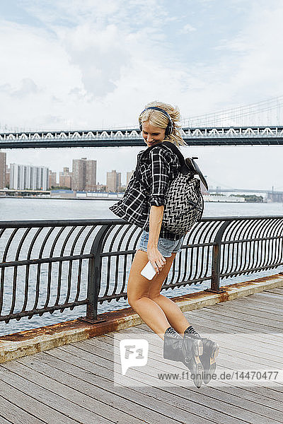 USA  New York City  Brooklyn  fröhliche junge Frau mit Kopfhörern und Handy am Wasser springend