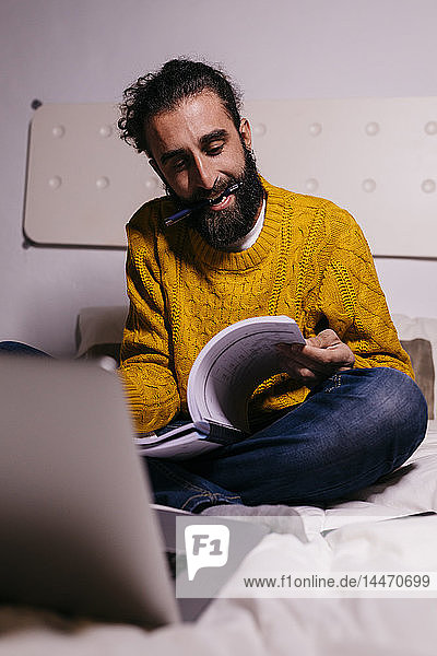 Young man working in bed at home with laptop and documents