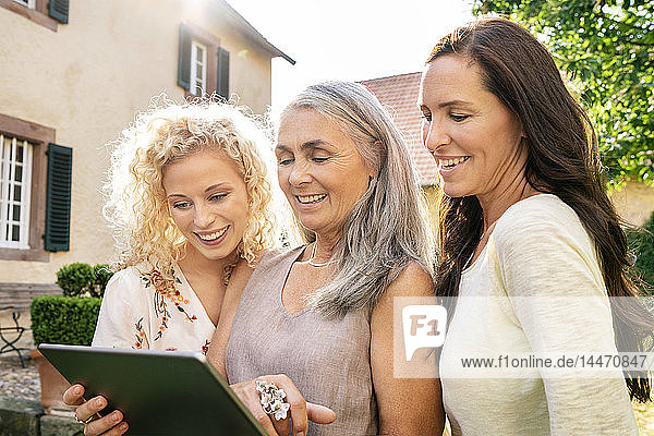 Drei lächelnde Frauen unterschiedlichen Alters verwenden Tablette im Garten