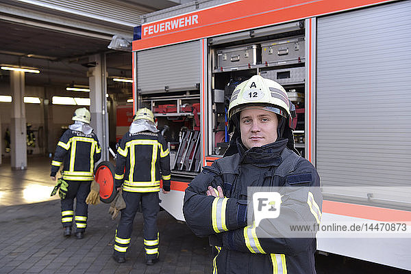 Porträt eines selbstbewussten Feuerwehrmannes vor dem Feuerwehrauto mit Kollegen im Hintergrund