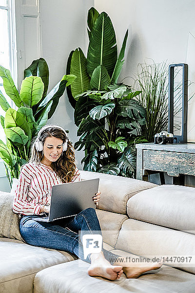 Lächelnde Frau mit Laptop und Kopfhörern auf der Couch sitzend