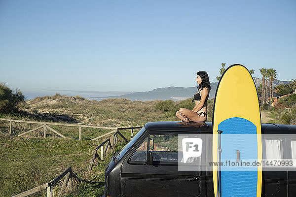Spanien  Andalusien  Tarifa  Frau auf dem Dach eines Wohnmobils sitzend  stehendes Paddelbrett an der Küste