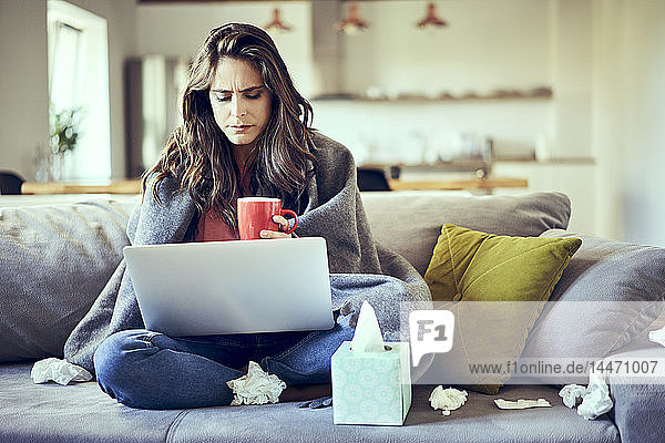 Eine kranke Frau sitzt auf einem mit einer Decke bedeckten Sofa mit einer Tasse Tee und einem Laptop