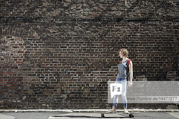 Junge Frau fährt mit einem Longboard an einer Ziegelmauer entlang