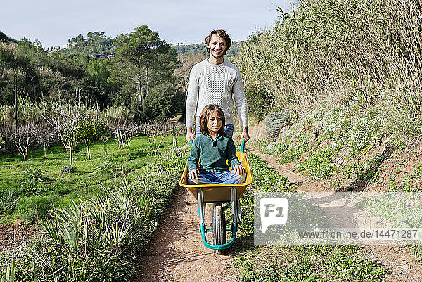 Vater zu Fuß auf einem Feldweg  Schubkarre schiebend  mit seinem Sohn darin sitzend