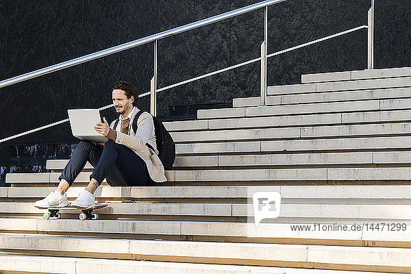 Man with backpack and skateboard sitting on stairs using laptop