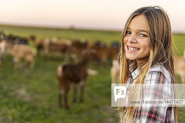 Porträt eines grinsenden Mädchens auf der Weide mit Ziegenherde im Hintergrund