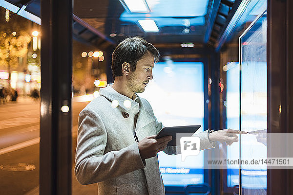 Geschäftsmann mit digitalem Tablett steht nachts an einer Bushaltestelle