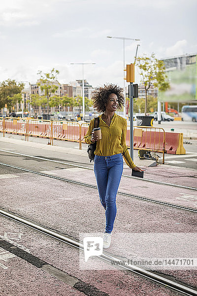 Lächelnde Frau mit Kaffee zum Mitnehmen und Notebook beim Überqueren von Gleisen in der Stadt