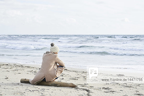 Spanien  Menorca  Rückenansicht einer älteren Frau mit Pudelmütze und Mantel  die im Winter am Strand sitzt