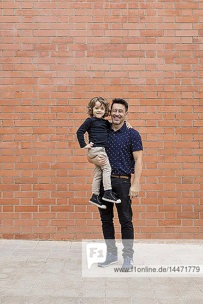 Porträt eines lächelnden Vaters  der seinen Sohn an einer Ziegelmauer trägt