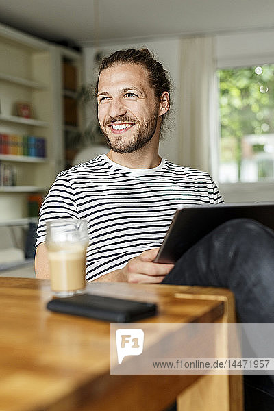 Junger Mann mit einem Brötchen zu Hause sitzend  mit digitalem Tablet