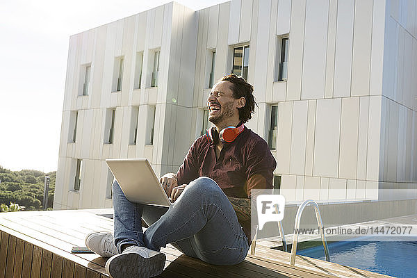 Mann sitzt im Schneidersitz auf einer Dachterrasse  benutzt Laptop  trägt Kopfhörer
