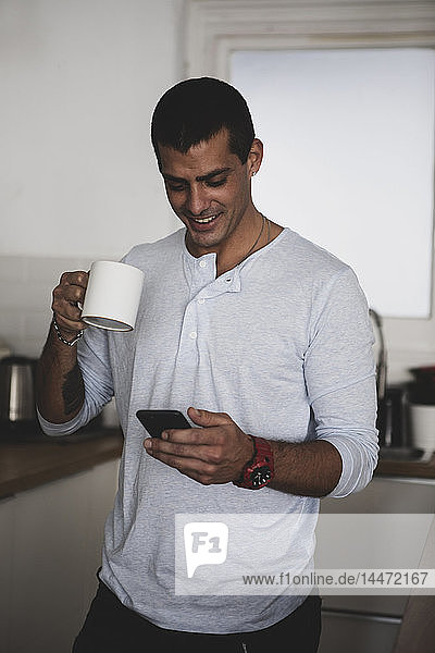 Lächelnder junger Mann mit einer Tasse Kaffee  der sein Handy in der Küche zu Hause benutzt