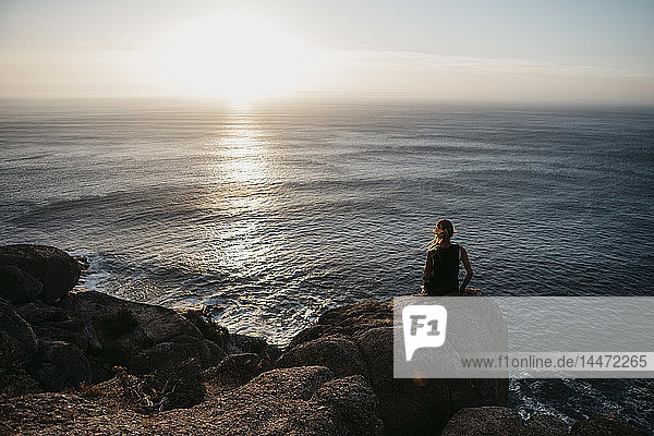Südafrika  Westkap  Frau sitzt auf einem Felsen und betrachtet den Sonnenuntergang  vom Chapman's Peak Drive aus gesehen