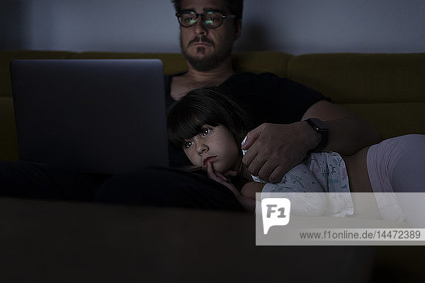 Father using laptop on couch at night with daughter watching