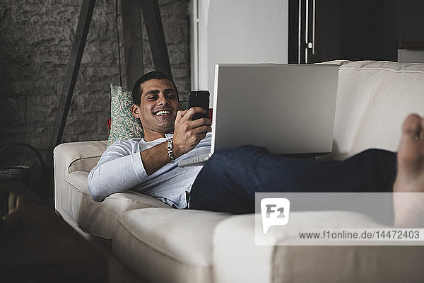 Glücklicher junger Mann liegt zu Hause auf dem Sofa und benutzt Handy und Laptop