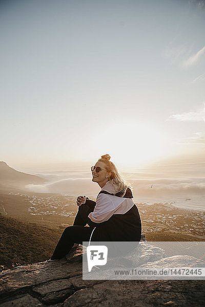 Südafrika  Kapstadt  Kloof Nek  lächelnde Frau  die bei Sonnenuntergang auf einem Felsen sitzt