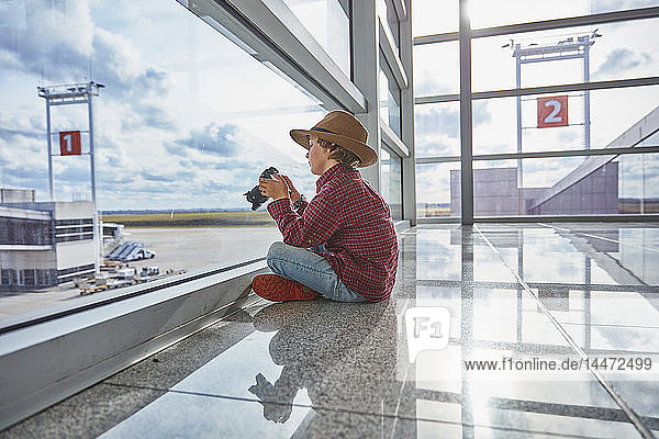 Boy sitting behind windowpane at the airport holding a camera
