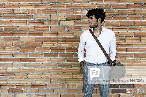 Junger Mann mit Tasche steht an Ziegelmauer und schaut sich um