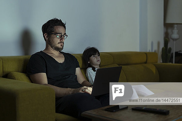 Father with daughter sitting on couch using laptop at night
