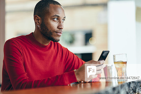 Pensive young man wearing red pullover at counter of a bar with soft drink and mobile phone