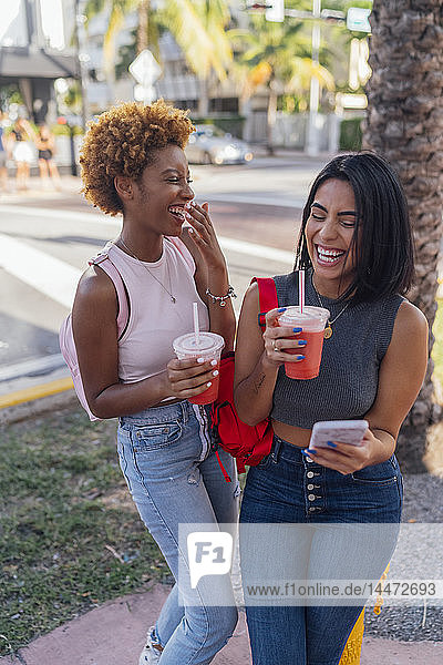 USA  Florida  Miami Beach  two happy female friends with cell phone and soft drink in the city