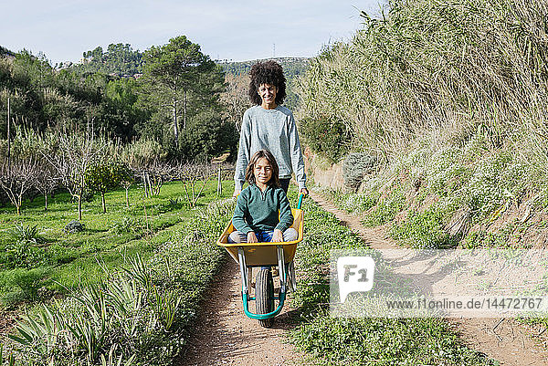 Mother walking on a dirt track  pushing wheelbarrow  with his son sitting in it