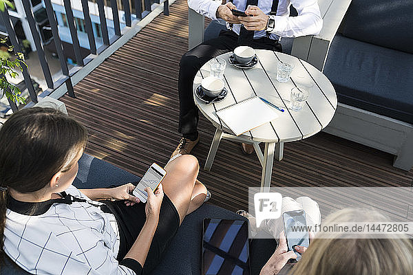 Three business colleagues having a coffee break on city terrace checking smartphones